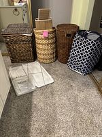 Five assorted hampers grouped on carpeted floor, including three wicker hampers, one fabric patterned hamper, and two white Ikea collapsible hampers