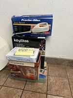 Photo of stacked boxed kitchen items showing Proctor Silex electric knife box and Keylitos hand blender box at kitchen floor level.
