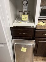 White KitchenAid stand mixer with stainless steel bowl and two large metal cooking sheets placed upright below the counter shelf.