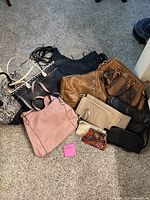 Multiple assorted purses of various styles and colors placed on carpeted floor.