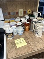 Wide view of 27 coffee mugs in different sizes and designs on kitchen counter near stove and outlet. Includes colorful and white mugs, some with text and logos.