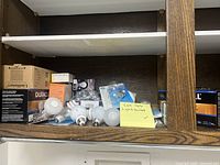 Wide view of open cabinet showing multiple boxes and loose light bulbs spread out on shelf, with note indicating light bulb lot