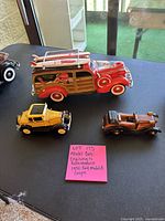 Three model cars: red Coca-Cola woody wagon with surfboards, yellow Ford Model A coupe, and small brown wood-effect model car on black surface.