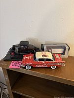 Three model cars displayed together on a wooden surface: 1931 Ford Model A Coupe, Coca-Cola Classic car, and boxed Freightliner M2 Van model.