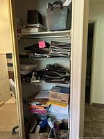 Metal shelf stocked with binders, notebooks, folders, pens, staplers, hole punches, tape, scissors, and other office supplies in boxes and containers.