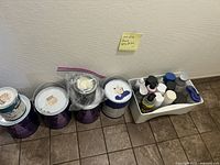 Box of assorted spray paint bottles and paint cans arranged on floor against white wall