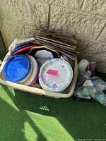 Overview photo showing a basket with paper plates, some with floral patterns, plain brown paper bags, plastic cups, cutlery, and straws on a balcony floor.