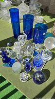 Photo showing frontage view of assorted paperweights, glass animal figurines including elephants, and three tall blue glass vases.