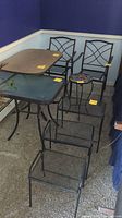 Two metal patio chairs with crisscross backs, rectangular and square glass top tables, flamingo glass top side table, and three stackable nesting tables arranged in a room corner.