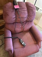 Full view of the power reclining chair upholstered in textured reddish fabric with cushioned back and armrests. Remote control with wire is placed on the chair.