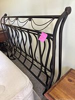 Side angle of black metal queen size headboard showing decorative twisted metal chains and curved vertical bars, placed against a wall next to mattress and wood nightstand.