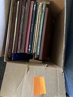 Side view of a cardboard box containing around 12 vinyl LP records with varied covers in browns and dark colors.