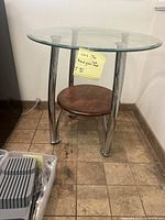 Full view of the modern round glass table showing the glass top, chrome legs, and wooden shelf underneath.