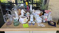 Overview of all items on a wooden table outdoors, showing pitchers, cups, vases, statues, and other knickknacks