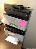 Samsung multifunction black and gray printer on top with scanning bed and control panel, stacked on two white Samsung printers.