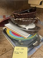 Stack of various serving trays and baskets showing round woven basket, wire basket, metal tray, white rectangular tray, colorful round tray, dark metal tray with handles
