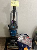 Full front view of upright Shark carpet cleaner with attached hose and bag of cleaning supplies on floor beside it.