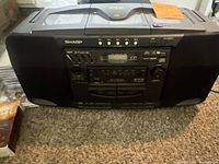 Photo showing the front view of Sharp black portable boombox with 5 CD changer on top, cassette deck and control buttons on front, placed on carpet.