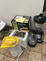 Photo showing two Presto PopLite popcorn makers with yellow plastic catchers and scoops, white toaster and blender with glass pitcher and black grinder attachment on kitchen floor.
