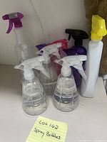 Spray bottles of various sizes and colors grouped together on shelf; includes clear plastic with white nozzles and taller bottles in purple, black, yellow, and white