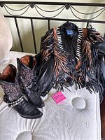 Photo showing the black leather-like fringe motorcycle jacket with brown and black fringes, white decorative elements, alongside a pair of black Tony Lama cowboy boots with boot helpers and embroidered details.