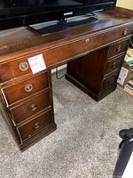 Photo showing full view of a medium to dark wood desk with multiple drawers, brass ring pulls, and a TV on top (TV not included). The floor is carpeted.