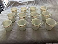 Twelve white opaque glass coffee mugs arranged in two rows on a cloth, showing all items included in the lot. The mugs have a consistent brown floral band design near the rim.
