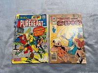 Two single issue comics on cloth background: Archie Captain Pureheart cover with hero fighting villain, showing wear; The Flintstones comic cover with characters and humor scene.