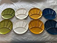 Eight ceramic fondue plates arranged in two rows showing colors green, white, mustard yellow, and dark blue with five segmented sections each.