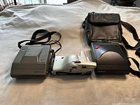 Group photo of three Polaroid cameras on a white cloth background, including one gray boxy model, one white JoyCam model, and one black instant camera with a carrying case.