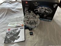 Overview of the assembled Millennium Falcon model with the box, instruction manual, stand, and tool laid out on a fabric surface.