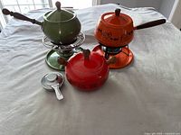 Photo showing the three colorful fondue sets arranged on a table: green set with pot on stand and burner, orange set with pot on stand and burner, red pot with lid and burner but no stand.