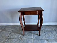 Front view of the wooden side table showing the drawer, missing knob, curved legs, and bottom shelf