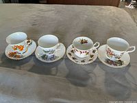 Four bone china teacups displayed with coordinating saucers on a soft surface, showing floral and maple leaf designs in varied colors.