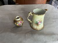 Photo of a vintage floral porcelain pitcher and a small vase on a grey surface, showing detailed hand-painted flowers and gold accents.