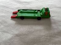 Top and side view of vintage green metal toy transporter truck with two red ramps at the rear, on white fabric background.