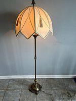 Full view of vintage floor lamp showing ornate metal base and illuminated fabric shade with tassels.