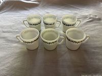 Six white Pyrex coffee mugs with floral patterns in green and blue arranged in two rows on a cloth surface