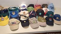 Wide view showing assortment of over 25 hats and visors in various colors and logos, laid out on a surface.