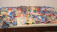 Multiple Disney VHS clamshell cover art sheets spread on table, showing collection size and variety