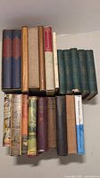 Top view of 16 assorted vintage books, some with dust jackets and sleeves, displayed upright and stacked in two rows under natural lighting.