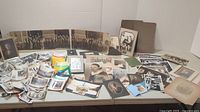 Display of a wide assortment of old black and white photographs laid out on a table including framed, loose, and folded photos.