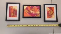 Three framed textile quilt art pieces hanging on a wall above a tape measure showing approximate size.