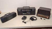 Photo showing a collection of vintage electronic devices including GE AM/FM cassette radio boombox, Sony Walkman cassette player, vintage General Electric brown wooden radio, headphones, and other devices.