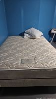 Full view of beige queen size mattress on box spring against blue wall, showing quilted pattern and pillows on top