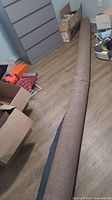 Carpet roll lying on wood floor among boxes and other items, showing full rolled length and texture of the carpet surface.