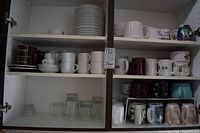 Cupboard shelves with stacks of white saucers, rows of white and coloured mugs, clear glasses on bottom shelf