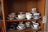 Multiple bone china tea cups and saucers on shelf, front and back rows