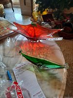 Two art glass pieces displayed side by side on a surface, showing one red-orange bowl-shaped glass and one green elongated curved piece with hanging crystals.