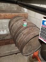 Wooden barrel stored on a basement floor near stairs. Barrel is medium to dark brown wood with six metal bands securing it. Shows signs of wear, rust, and aging.